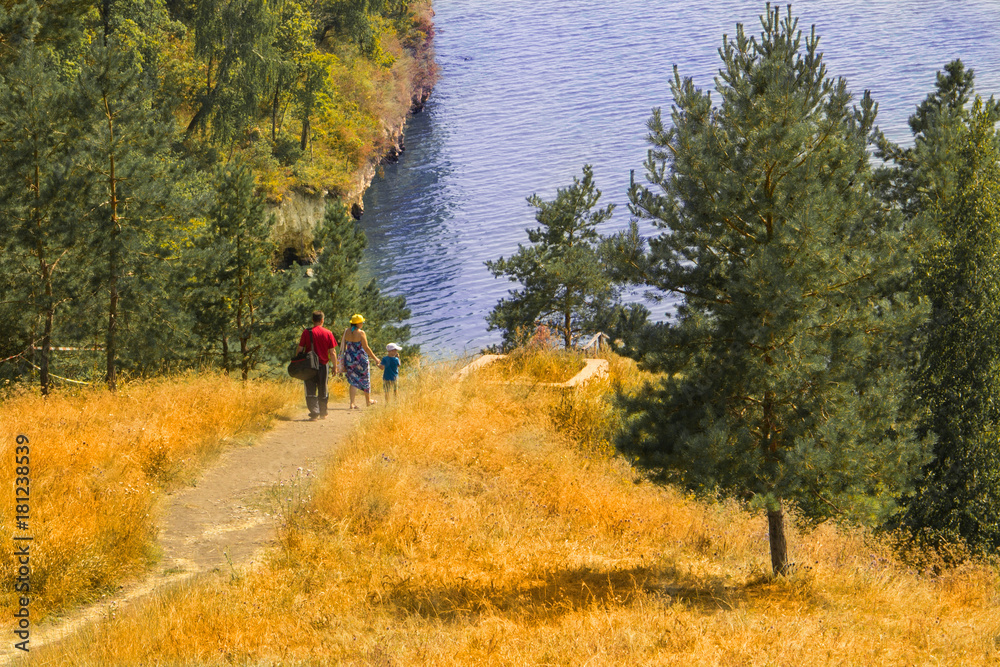 A man with a bag, a woman in a dress, a hat and a bag and a child in shorts, a T-shirt and a cap walking along a sand trail among the yellow grass and green trees to the river bank.