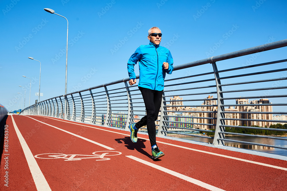 Mature sportsman in activewear jogging along bridge over river and cityscape