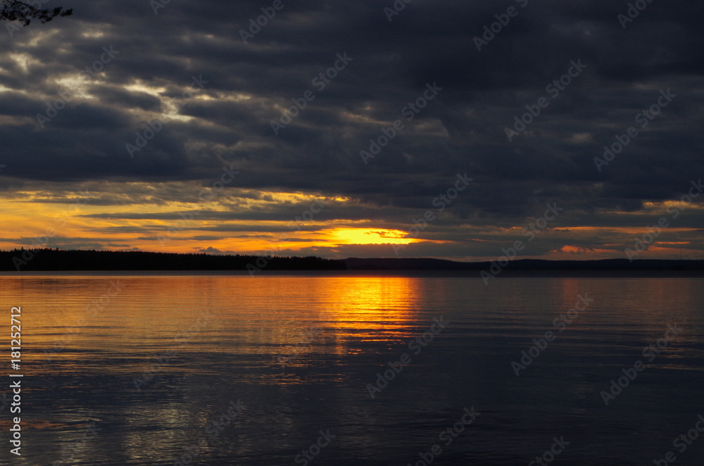 Sunset sky panorama. Bright sun line against dark clouds background. Reflection on a water.