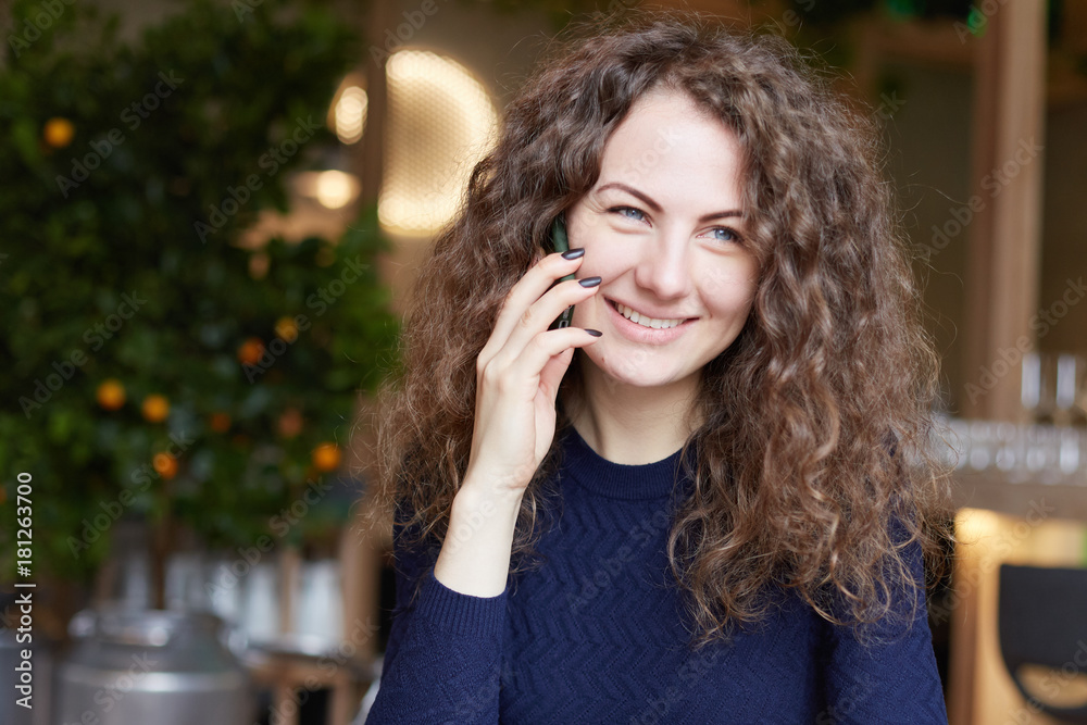 Indoor shot of stylish attractive woman with curly hair in blue sweater, calls her friend over smart phone, sits in restaurant, smiling charmlike, looks at window. Technology and communication. Indoor shot of stylish attractive woman with curly hair in blue sweater, calls her friend over smart phone, sits in restaurant, smiling charmlike, looks at window. Technology and communication.