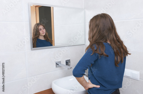 Beautiful teenage girl touching her face before the mirror, she found acne problem, standing in a bathroom and looking at her reflection in a mirror.