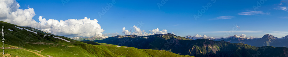 Fototapeta premium Wide mountain panorama.Green hills with snow on peaks in evening sunlight. Dark shadows. Blue sky with some clouds. Summer in Georgia.