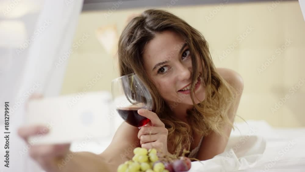 Attractive woman drinks red wine and takes selfie on her smartphone on bed Attractive woman drinks red wine and takes selfie on her smartphone on bed
