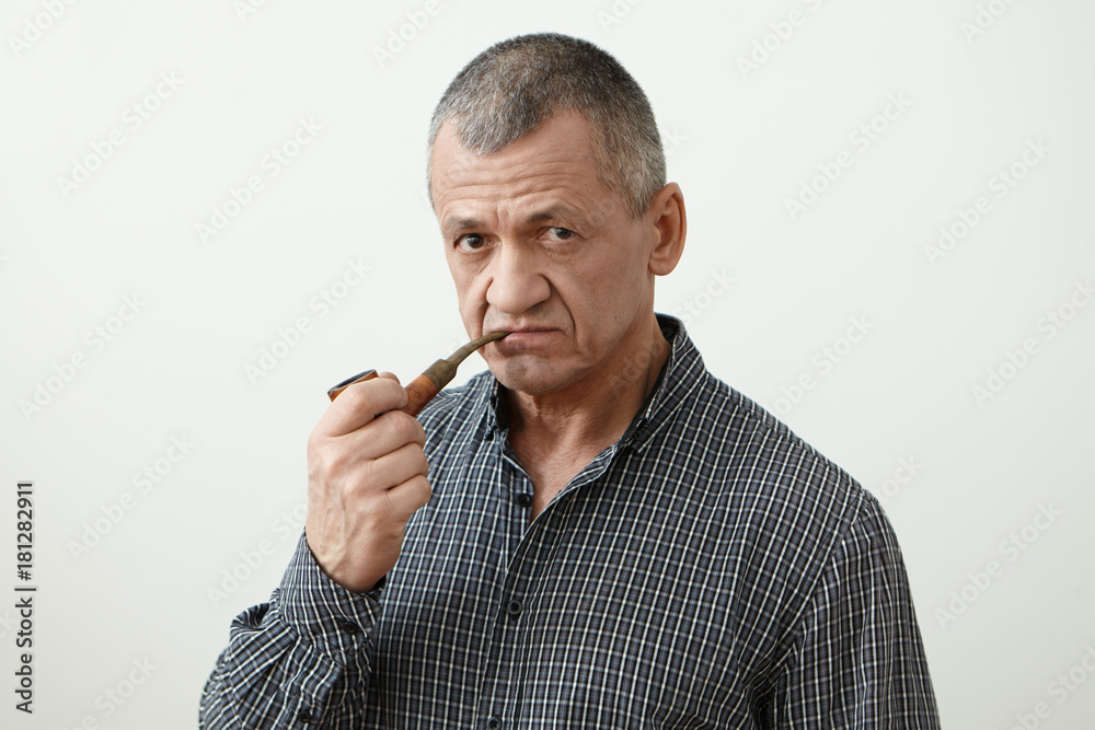 Isolated portrait of serious senior male with grey hair posing in studio, smoking pipe and looking at camera with thoughtful expression. Elderly man wearing shirt enjoying tobacco pipe after dinner Isolated portrait of serious senior male with grey hair posing in studio, smoking pipe and looking at camera with thoughtful expression. Elderly man wearing shirt enjoying tobacco pipe after dinner