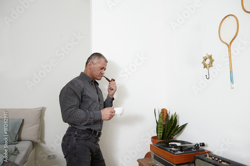 Stylish senior man wearing shirt and jeans standing in modern apratment interior hoding cup of coffee, smoking tobacco pipe and listening to music on retro vinyl record player, deep in thoughts photo