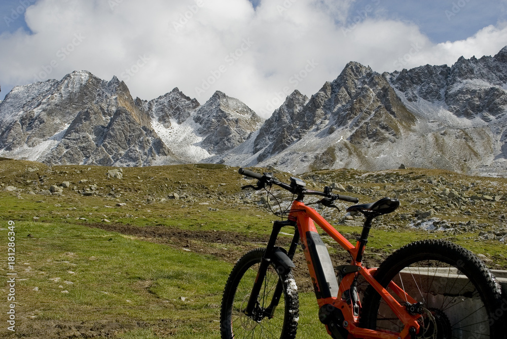 electric bicycle (orange), ebike, in a valley covered with pasture grass, snow covered mountains background (Castel, Kastelhorn) from a fresh snowfall, Formazza valley, Autumn, clouds sun, Italy