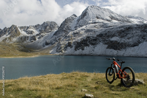 electric bicycle (orange), ebike, alpine lake with green water, in background snow covered mountains (Castel, Kastelhorn) from a fresh snowfall, Formazza valley, Autumn, clouds sun, Piedmont, Italy