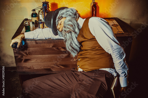 Bearded man with the bottle in one hand is sleeping at the table.