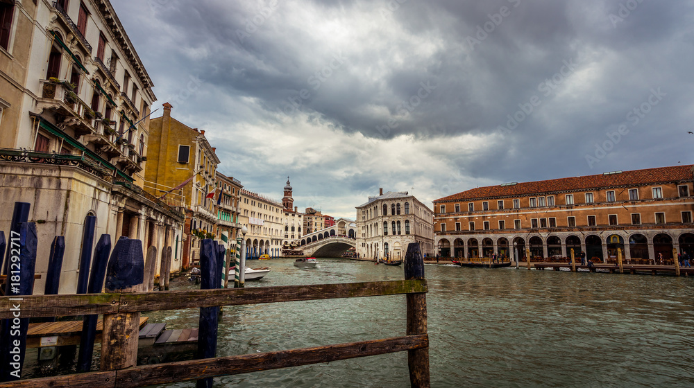 Panoramic view on famous Grand Canal among historic houses in Venice, Italy at cloudy day with dramatic sky, wood bridge on the foreground an Rialto bridge on the background