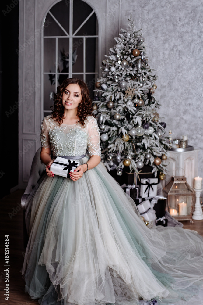 The young woman the brunette with curly hair in an elegant blue dress against the background of the Christmas fir-tree. The girl smiles, holds a New Year's gift in hand and looks in a shot.Portrait to