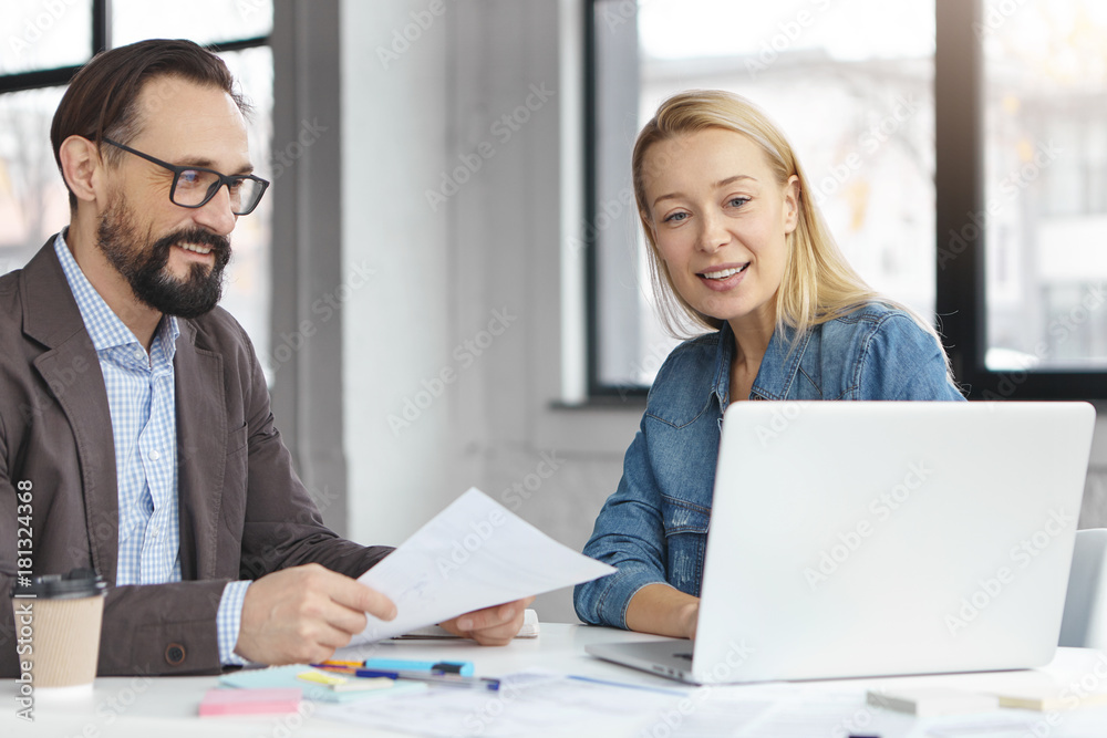 Experienced employees look at computer monitor, watch presentation of marketeer, work on business papers. Meeting for brainstroming. Blonde female and bearded man speak about startup at office Experienced employees look at computer monitor, watch presentation of marketeer, work on business papers. Meeting for brainstroming. Blonde female and bearded man speak about startup at office