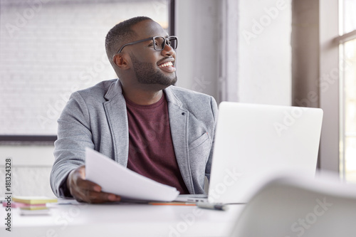 Happy dark skined male manager wears classic suit, works on computer and with documents, sits at spacious cabinet, makes banking online. Cheerful middle aged African male owner of business company