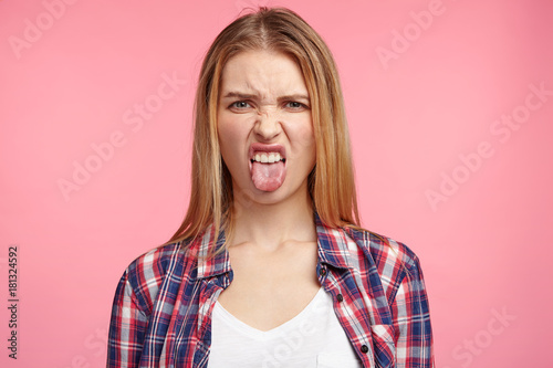 Dissatisfied female model frowns face, has disgusting expression, shows tongue, expresses non compliance, irritated with somebody, rejects do something. People and negative facial expressions photo