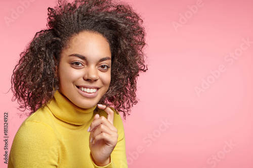 Cheerful dark skinned female with bushy Afro hairstyle, wears yellow poloneck sweater, happy to be photographed in popular magazine on cover page, being famous model, stands over pink background