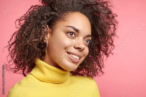 Close up portrait of pretty smiling African female has dark pure healthy skin, looks pensively aside, thinks or remembers pleasant moments in life, dreams about buying new apartment, poses at stusio photo