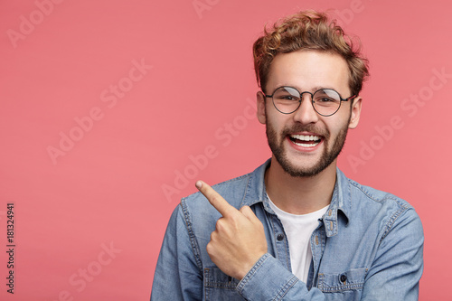 Stylish unshaven man in denim shirt points at copy space on pink wall as shows something pleasant, has smiling look, advertises product. Look over here! People, advertising, emotions concept