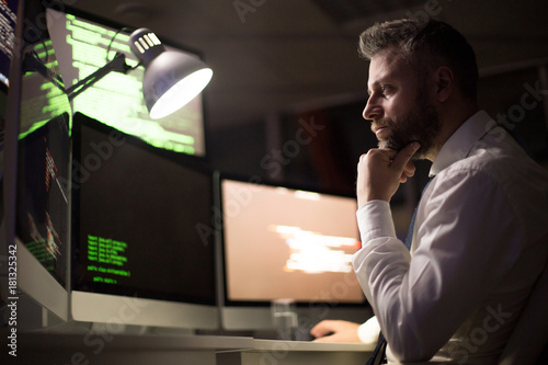 Profile view of pensive bearded coder wrapped up in work while sitting in front of modern computer, interior of dim office on background