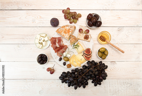 Overhead of cheese plate with pieces moldy cheese, prosciutto, pickled plums, olives, grapes, figs, honey, pear, nuts on white wooden background. Top view. photo