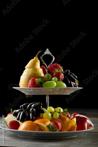 Vertical shot of various fruits served on a silver plate on black background sliced pear strawberries grapes orange apples vitamins health healthy eating dieting serving concept.