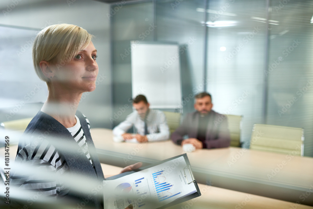 Portrait of ambitious young woman standing in office looking away hopefully and smiling with three people at meeting table in background, copy space - obrazy, fototapety, plakaty 