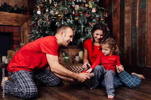 family in pajamas plays next to a Christmas tree and a fireplace. A little daughter takes a gift from her father.