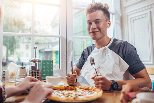 Obraz na plátně Stylish teenager wearing napkin holding knife and fork ready to eat pizza sittin
