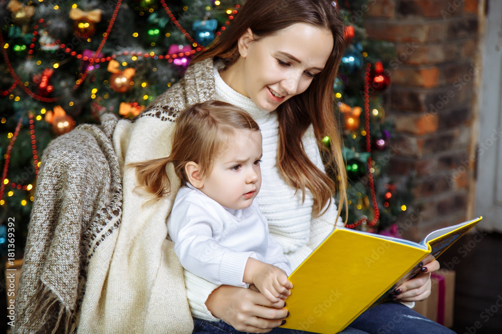 Pretty young mom reading a book to her cute daughter near Christmas tree indoors.