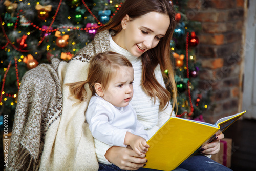 Pretty young mom reading a book to her cute daughter near Christmas tree indoors.