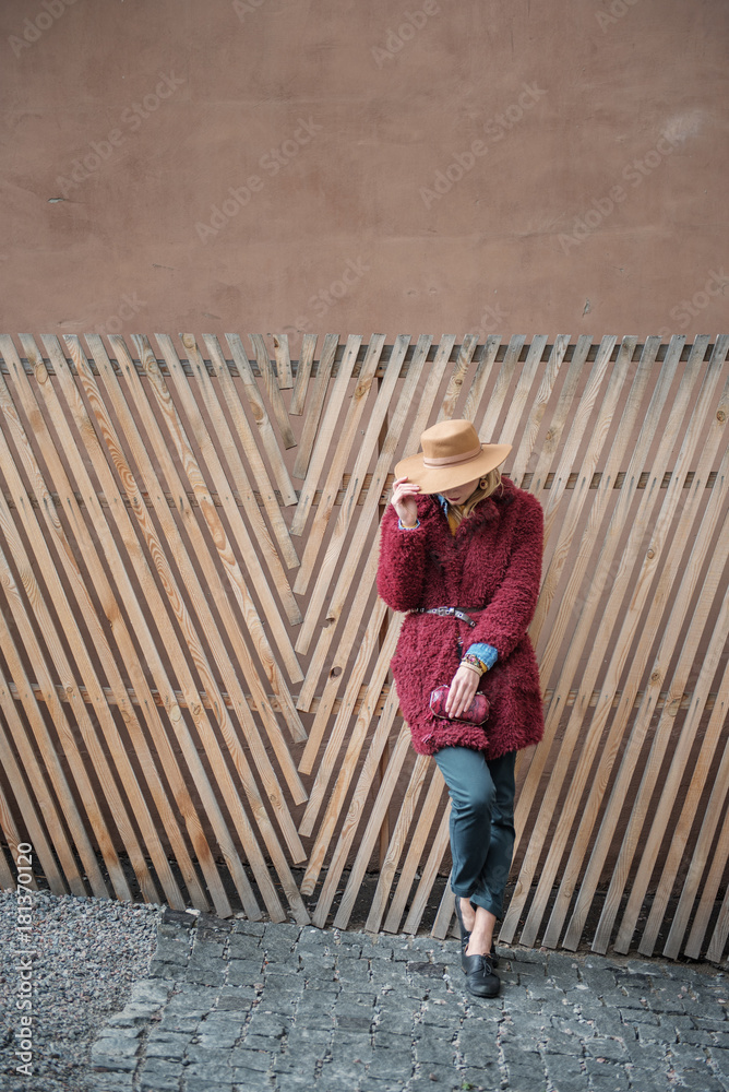 Full length of glamorous blond girl standing near wood fence while hiding face by trendy hat. She is carrying small purse. Copy space Full length of glamorous blond girl standing near wood fence while hiding face by trendy hat. She is carrying small purse. Copy space