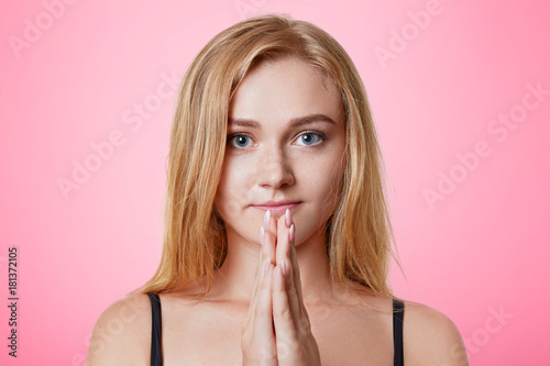 Murais de parede Gorgeous light haired female keeps palms together, has faithful expression, prays for good luck or fortune, looks directly into camera, isolated over pink background