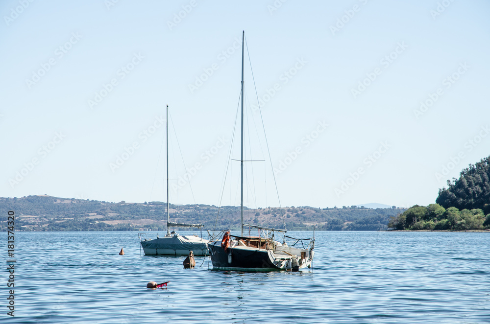 Two sailboats anchored in the lake. In the background campgrounds and hills with woods.