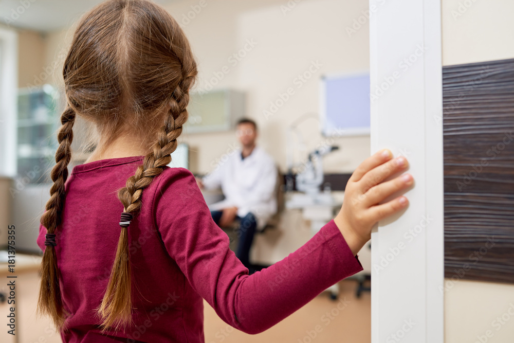 Back view portrait of scared little girl entering doctors office, standing in doorway and looking at blurred pediatrician Back view portrait of scared little girl entering doctors office, standing in doorway and looking at blurred pediatrician