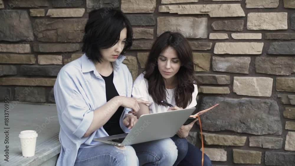 Two young and beautiful women are sitting on a porch and working together. One is holding a laptop, the second is writing. Handheld real time establishing shot Two young and beautiful women are sitting on a porch and working together. One is holding a laptop, the second is writing. Handheld real time establishing shot