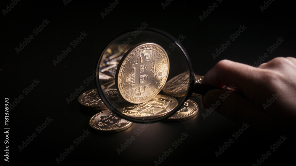 Fototapeta premium Unrecognizable person holding magnifying glass and looking through it on new virtual money golden bitcoin placed on a black background.