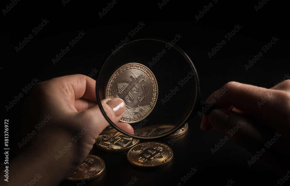 Fototapeta premium Unrecognizable person holding magnifying glass and looking through it on new virtual money golden bitcoin placed on a black background.