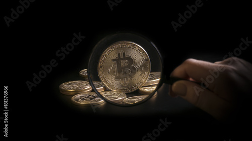 Unrecognizable person holding magnifying glass and looking through it on new virtual money golden bitcoin placed on a black background.