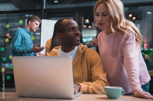 Helpful device. Low angle of african man is looking with smile at female colleague while sitting at table with laptop. She is leaning on desk and gazing at screen with concentration