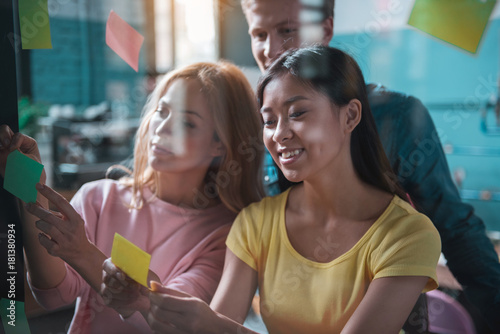 Portrait of beaming asian and caucasian girls labeling post-it notes on window in modern office. Man looking at it. Creativity concept