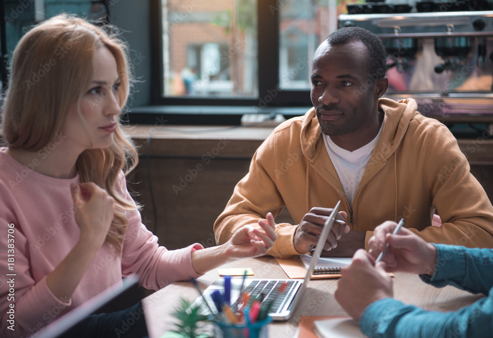 Portrait of orderly african male looking at colleague while sitting at table. Serene caucasian woman flourishing arms while telling with associate. Discussion concept