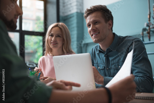 Low angle portrait of beaming young girl and happy man telling plan to their colleague while locating at table. Startup team concept