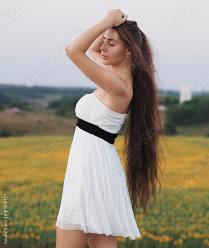 Young brunette model at the white dress staying near the sunflowers field and looking forward
