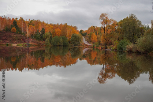 The autumn forest and the road on the hills are reflected in the calm water of the forest lake. © NATALIA