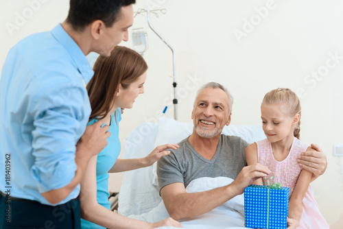 An elderly man lies in a hospital room on a bed. He is seen by a man with a woman and granddaughter.