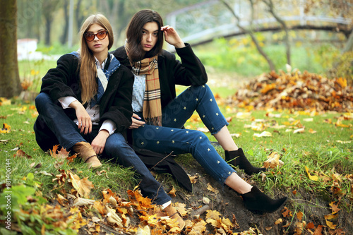 Outdoors lifestyle fashion portrait of two sexy girls friends, posing on the autumn park. Wearing stylish bright outerwear and sunglasses. Autumn colors
