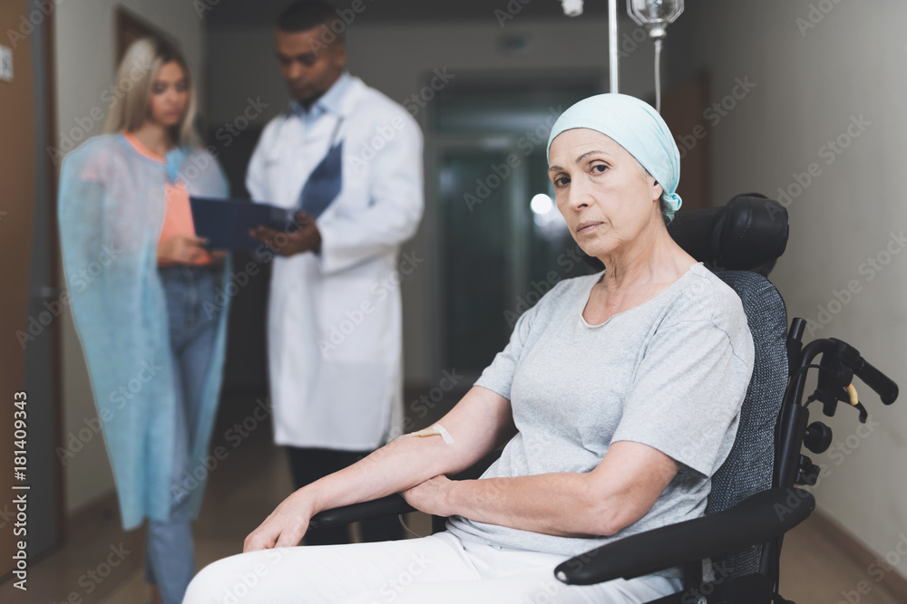 Obraz premium A woman with cancer, with a doomed look, is sitting in a wheelchair. Behind her daughter stands and talks to the doctor.
