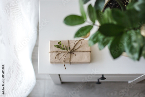 gift wrapped in kraft paper tied with string decorated with a branch with green leaves lies on a white bedside table