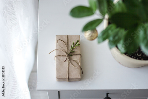 A gift wrapped in kraft paper tied with a rope lies on a white wooden bedside table next to a flower in a pot decorated for Christmas near the window