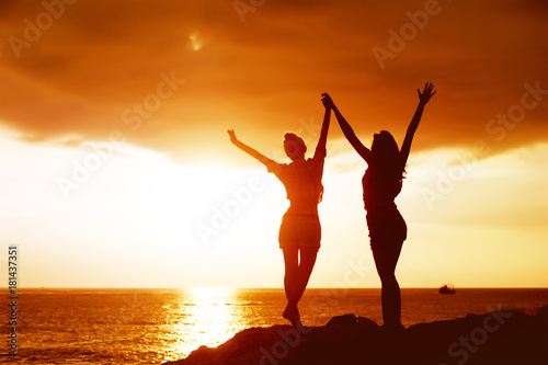 Two happy girls with raised arms on background of sunset sea