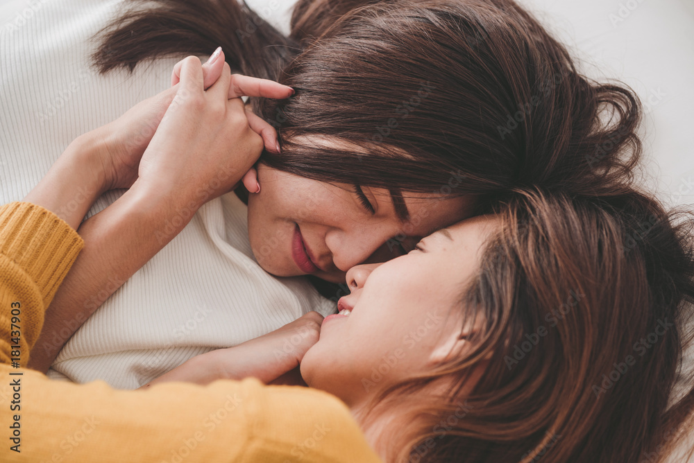 Fototapeta premium Top view of beautiful young asian women lesbian happy couple hugging and smiling while lying together in bed under blanket at home. Funny women after wake up. Lesbian couple together indoors concept.