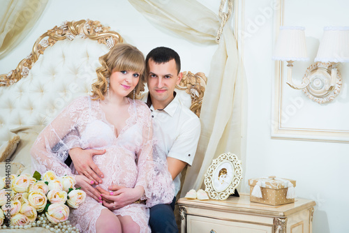 Beautiful couple, pregnant young woman and man hugging with love kisses, sitting on the bed, in a home interior.